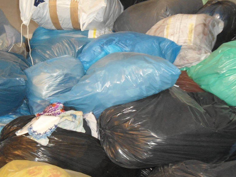 Stos worków z odzieżą, w tym niebieskie, czarne i białe, niektóre z widoczną zawartością, tworzące chaotyczny, ale uporządkowany wizualnie stos.