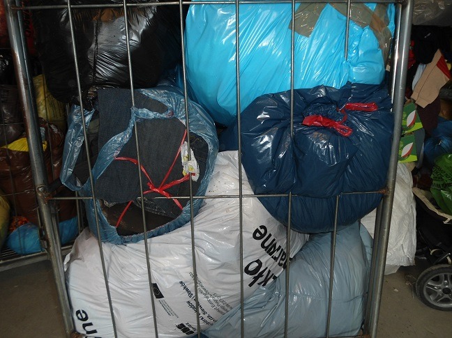 Metalowy kosz wypełniony workami z odzieżą używaną w różnych kolorach: niebieski, czarny, biały. Worki są szczelnie zamknięte i gotowe do transportu.