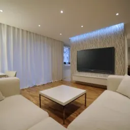 Nowoczesny salon z drewnianą podłogą, białymi sofami, stolikiem kawowym i telewizorem na ścianie z podświetleniem LED, widok na biurko z lampą i zasłonięte okno.