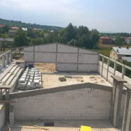 Budowa hali z betonu komórkowego w stanie surowym otwartym, widok z góry na wznoszone ściany, strop i przygotowane bloczki, w tle wiejski krajobraz.