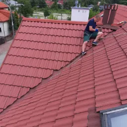 Pracownik na dachu z czerwonej dachówki, zabezpieczony liną, obok komina i okna dachowego, w tle zabudowa jednorodzinna.