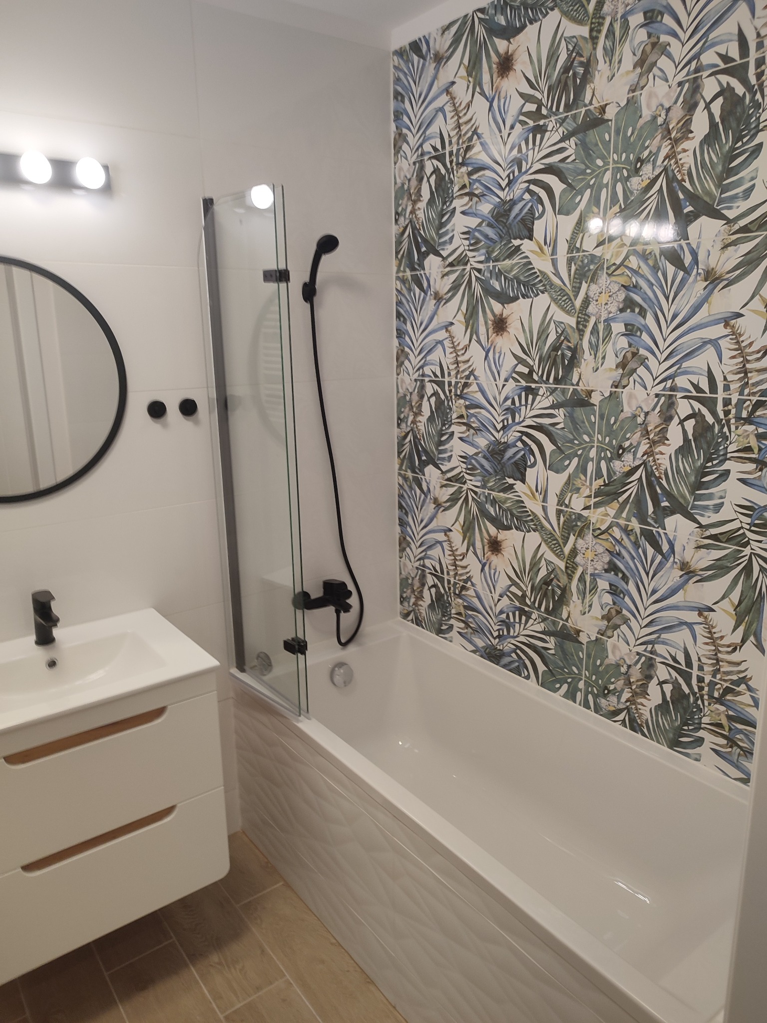 Nowoczesna łazienka z białą armaturą, kabiną prysznicową z czarnymi elementami i ścianą w płytkach z motywem tropikalnych liści.