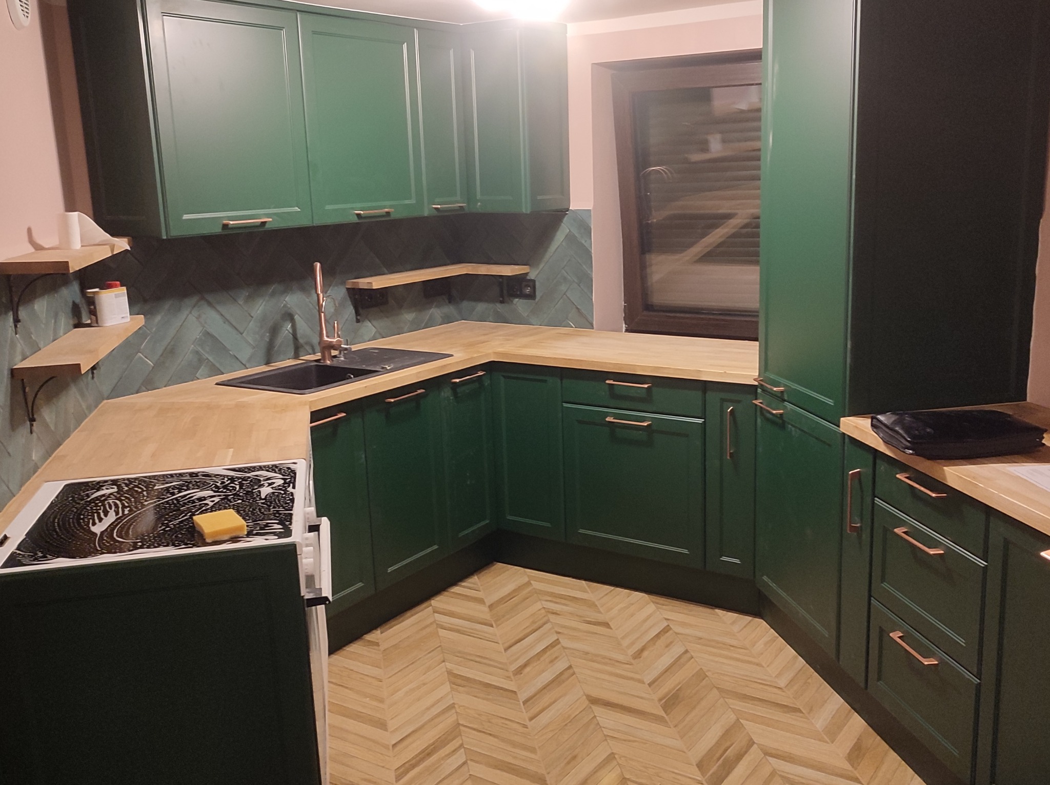 Nowoczesna kuchnia z zielonymi szafkami, drewnianym blatem i podłogą ułożoną w jodełkę; na płycie indukcyjnej gąbka do mycia naczyń.