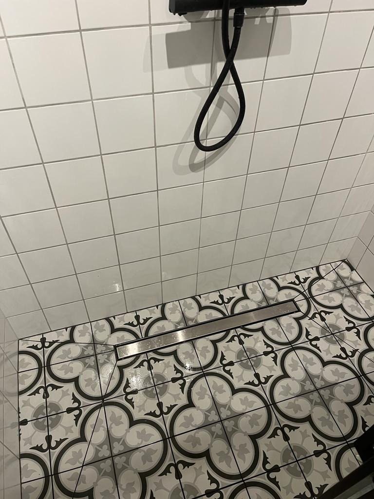 Nowoczesna kabina prysznicowa z białymi, kwadratowymi płytkami ściennymi i podłogą wyłożoną dekoracyjnymi płytkami w odcieniach szarości i czerni z motywem czterolistnej koniczyny. Widoczny liniowy...