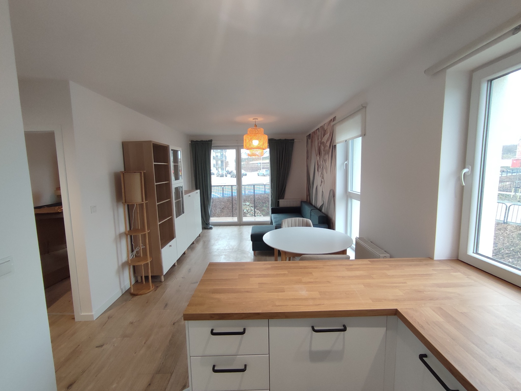 Jasne, przestronne wnętrze mieszkania z drewnianą podłogą, widokiem na balkon i nowoczesną zabudową kuchenną z drewnianym blatem, oświetlone oryginalną lampą z abażurem.