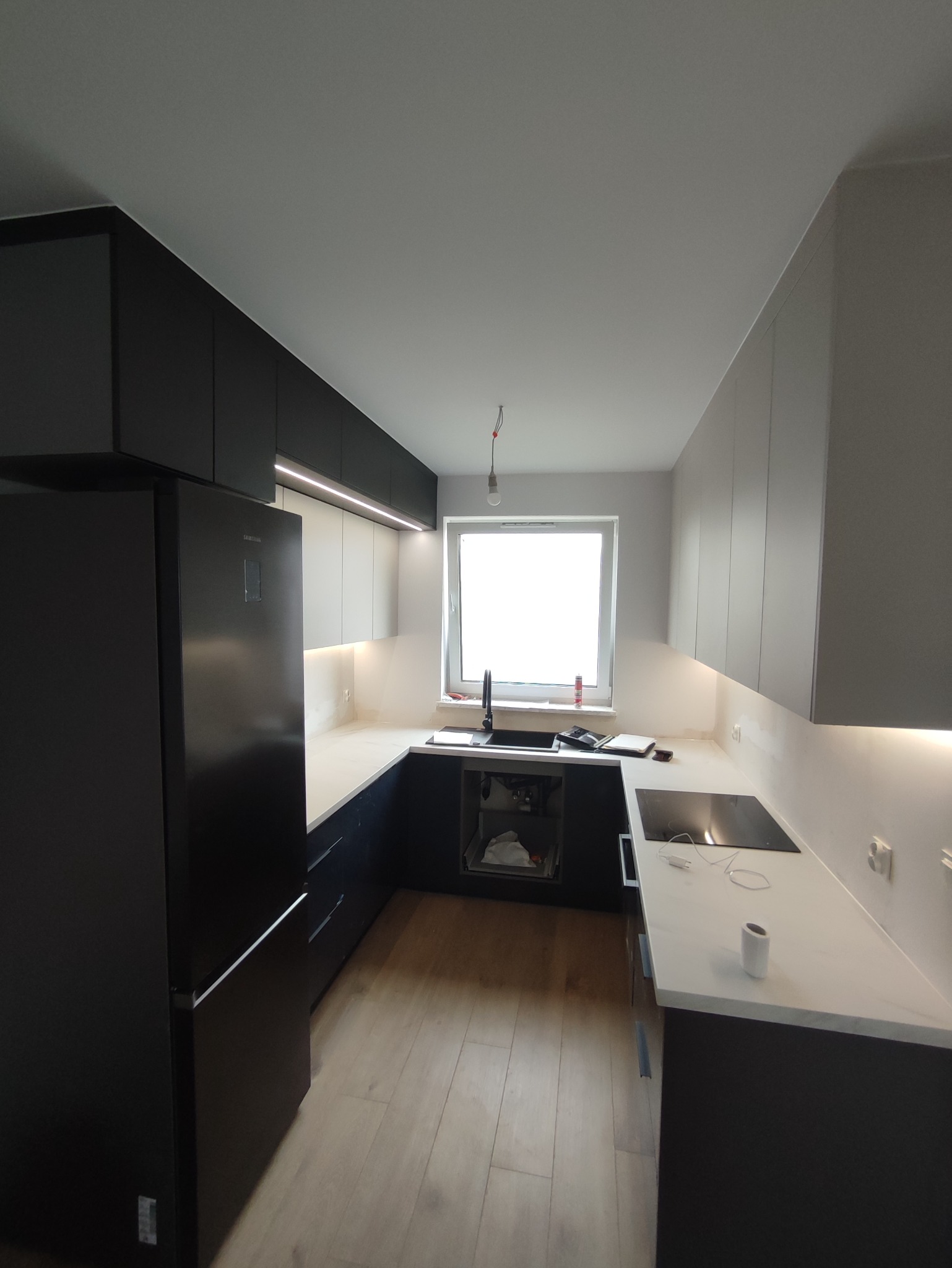 Nowoczesna kuchnia w trakcie wykańczania: czarno-białe szafki, biały blat, czarny zlew, płyta indukcyjna, oświetlenie LED pod szafkami, widoczne przewody elektryczne.