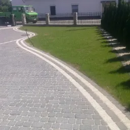 Ułożona z szarych kostek brukowych wijąca się ścieżka z jasnymi obrzeżami, prowadząca do bramy z metalowym ogrodzeniem i kamiennymi słupkami. Obok trawnik z rzędem tuj. W tle zielony samochód...