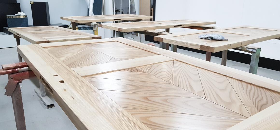 Kilka niedokończonych, drewnianych drzwi z wzorem w jodełkę, leżących na kozłach malarskich w warsztacie.