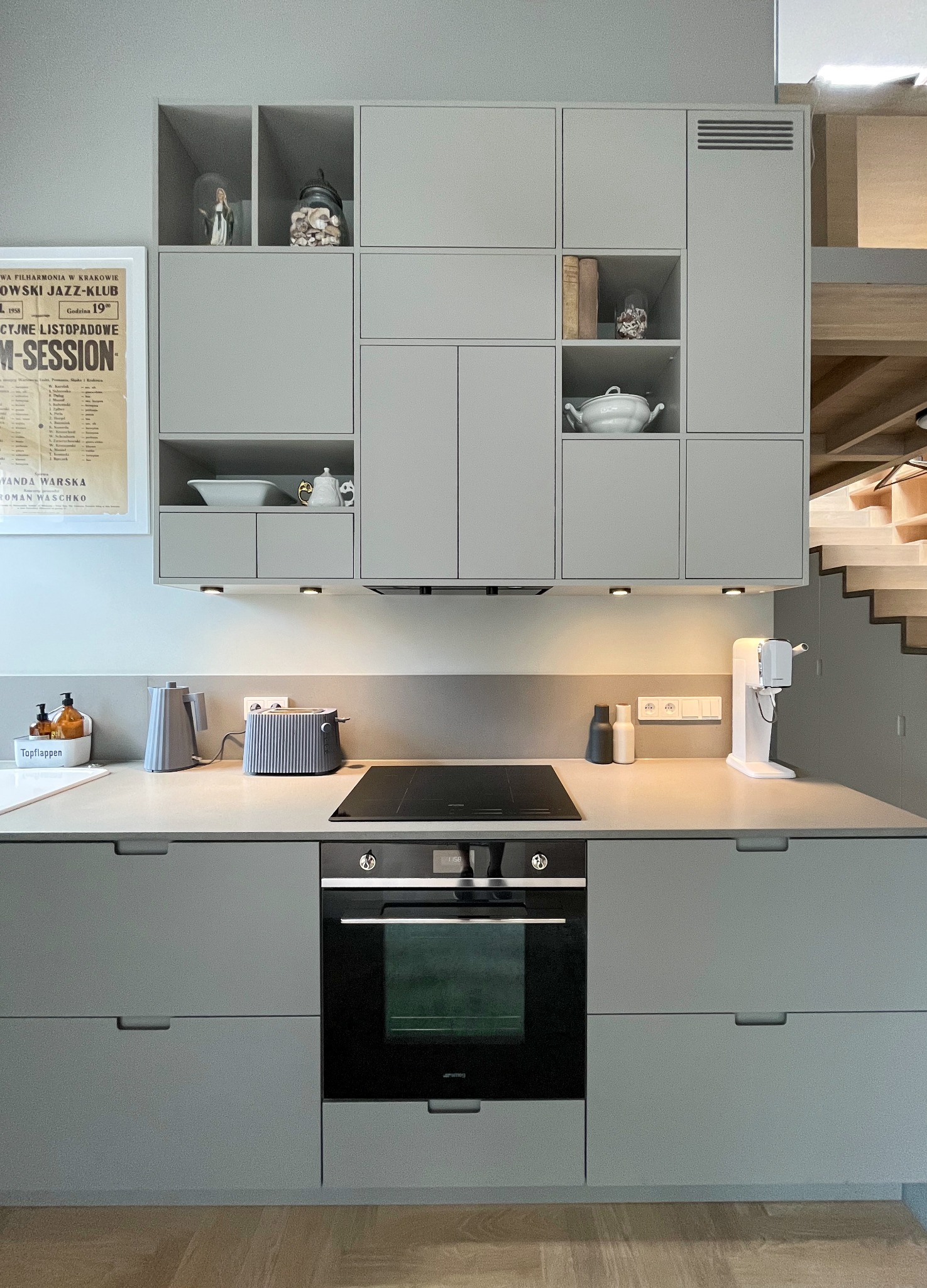 Szare, minimalistyczne meble kuchenne z piekarnikiem Smeg, płytą indukcyjną, czajnikiem, tosterem i ekspresem do wody sodowej. Na półkach dekoracje i plakat z Krakowskiego Jazz-Klubu.