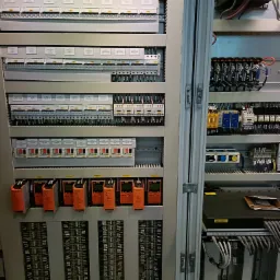 Rozdzielnica elektryczna z widocznymi bezpiecznikami, przekaźnikami, sterownikami PLC i okablowaniem w szafie sterowniczej, widoczne oznaczenia i etykiety.