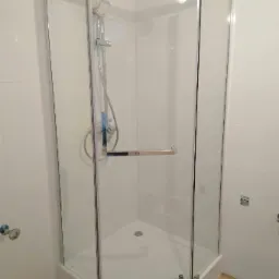 Nowoczesna, narożna kabina prysznicowa z chromowanymi okuciami i deszczownicą, osadzona na białym brodziku, w łazience z jasnymi płytkami i drewnopodobną podłogą.