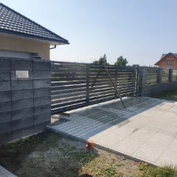 Nowoczesna brama przesuwna z poziomych profili, zamontowana na słupkach z szarej kostki łupanej. Widoczne płytki chodnikowe i fragment domu w tle.