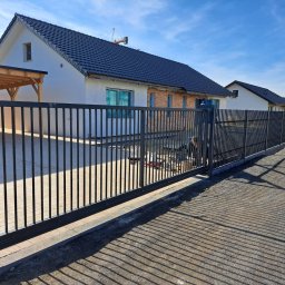 SIEDY PAWEŁ BETOMET - Nowoczesna, przesuwna brama wjazdowa z metalowymi sztachetami na tle domu jednorodzinnego w trakcie budowy. Widoczne narzędzia i osoba montująca.
