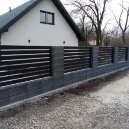 Nowoczesne ogrodzenie z szarej kostki łupanej i czarnych, poziomych paneli na tle domu z ciemnoszarą dachówką i drzew bez liści.