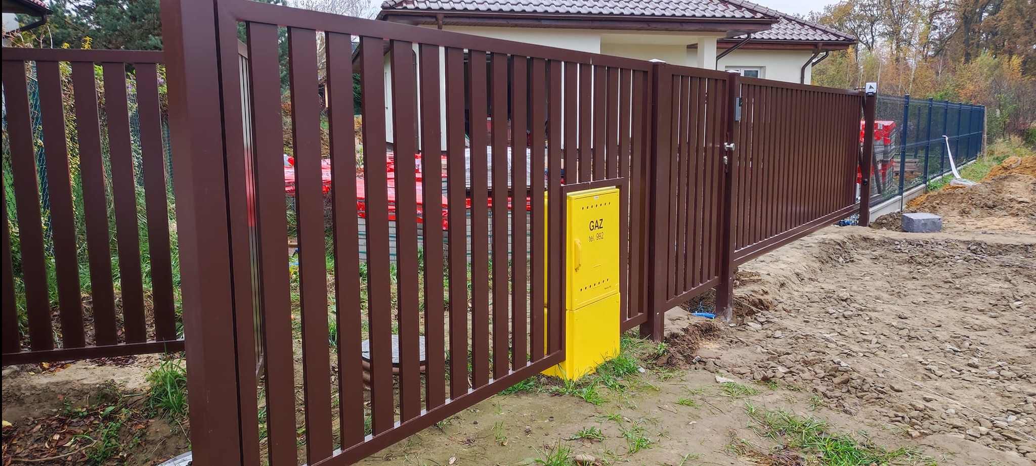Nowoczesna brama wjazdowa z brązowych, pionowych paneli metalowych, z żółtą skrzynką gazową po lewej stronie, na tle domu z brązowym dachem i częściowo widocznym ogrodzeniem siatkowym.