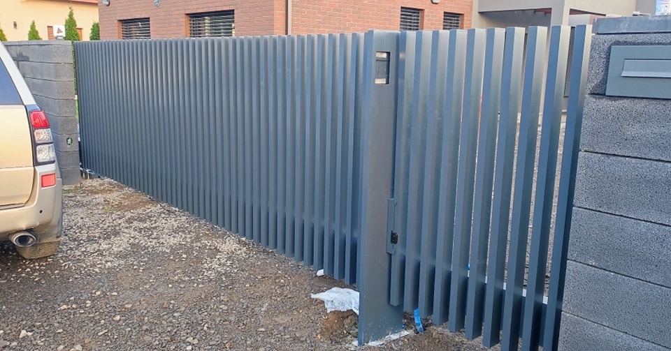 Nowoczesna, szara brama wjazdowa z pionowych, metalowych profili, zamontowana między betonowymi słupkami z wbudowaną skrzynką na listy, widok z zewnątrz posesji.