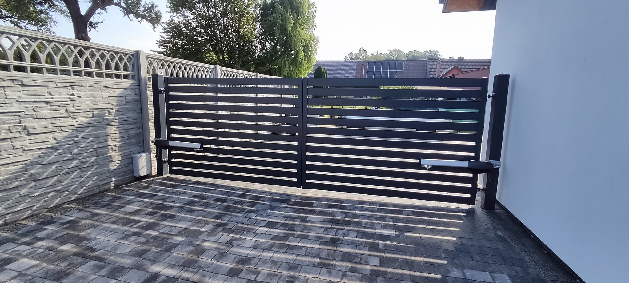 Nowoczesna, pozioma brama wjazdowa z automatyką, zamontowana między ogrodzeniem z paneli imitujących kamień a białą ścianą budynku. Ciemne deski bramy kontrastują z jasną kostką brukową...