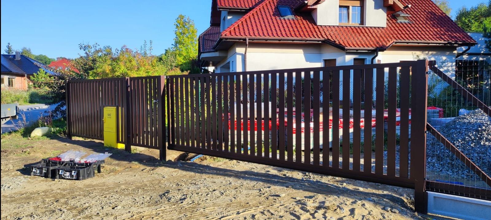 Nowoczesna, brązowa brama przesuwna z pionowych profili, widoczna skrzynka gazowa w żółtym kolorze i narzędzia montażowe w pobliżu, dom z czerwoną dachówką w tle.