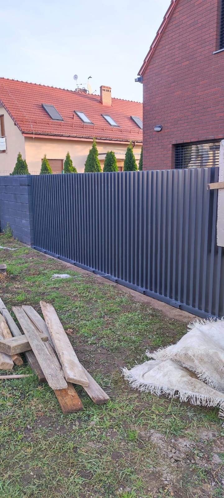 Nowoczesne, grafitowe ogrodzenie metalowe z pionowych profili, zakończone betonowymi słupkami, na tle domu z czerwonej cegły i zielonych tuj. Na trawie leżą deski i zabrudzona plandeka.