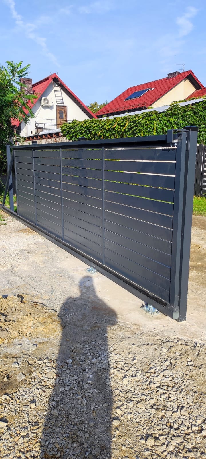 Nowoczesna, przesuwna brama wjazdowa z poziomymi panelami w kolorze grafitowym, zamontowana na betonowej podstawie, widoczny cień fotografa.