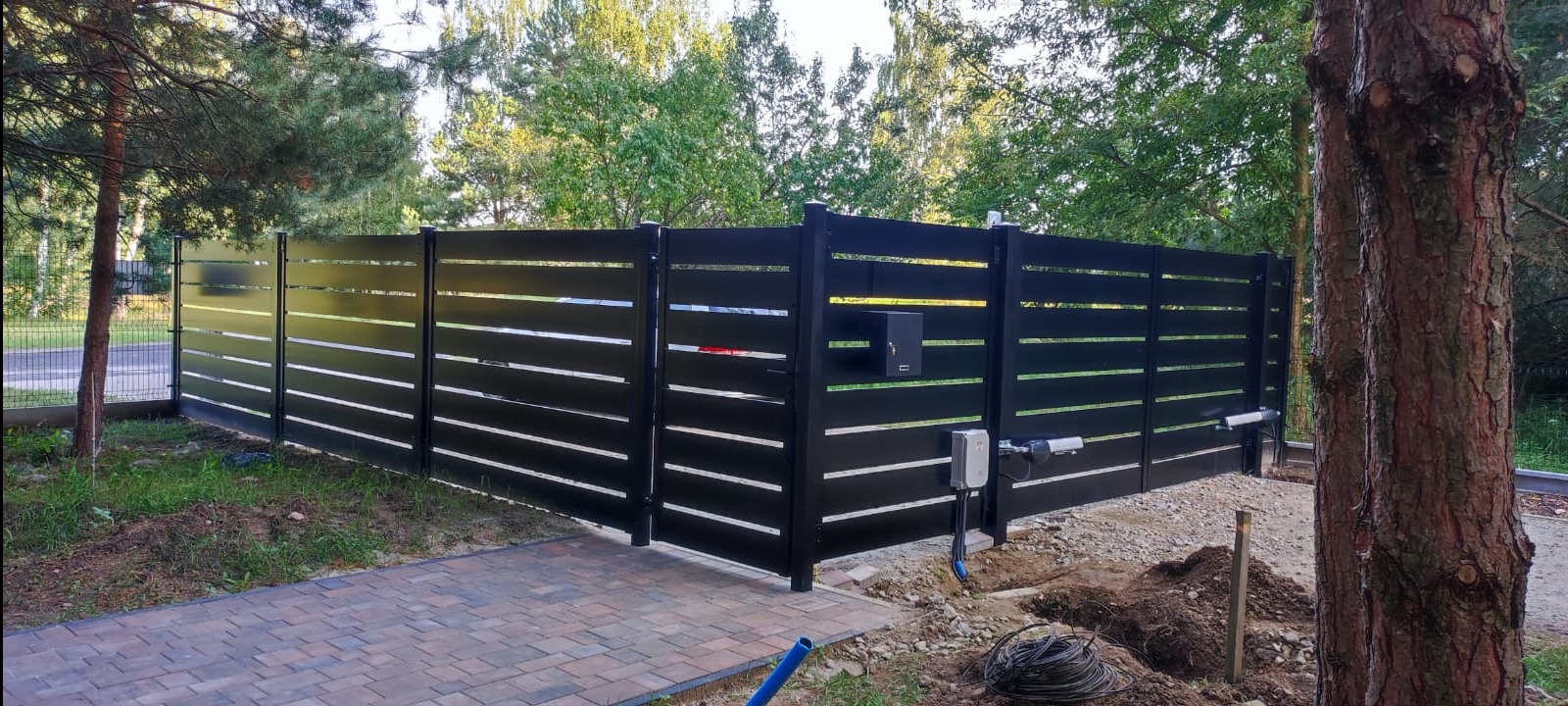 Nowoczesne, czarne ogrodzenie panelowe z poziomymi szczelinami i automatyczną bramą wjazdową, widoczne elementy montażowe i okablowanie, kostka brukowa prowadząca do bramy, drzewa w tle.