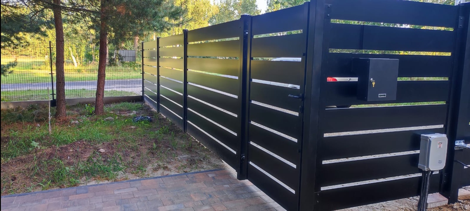 Nowoczesne, czarne ogrodzenie z poziomymi panelami i skrzynką na listy, widoczna automatyka do bramy, kostka brukowa u podnóża, zieleń w tle.