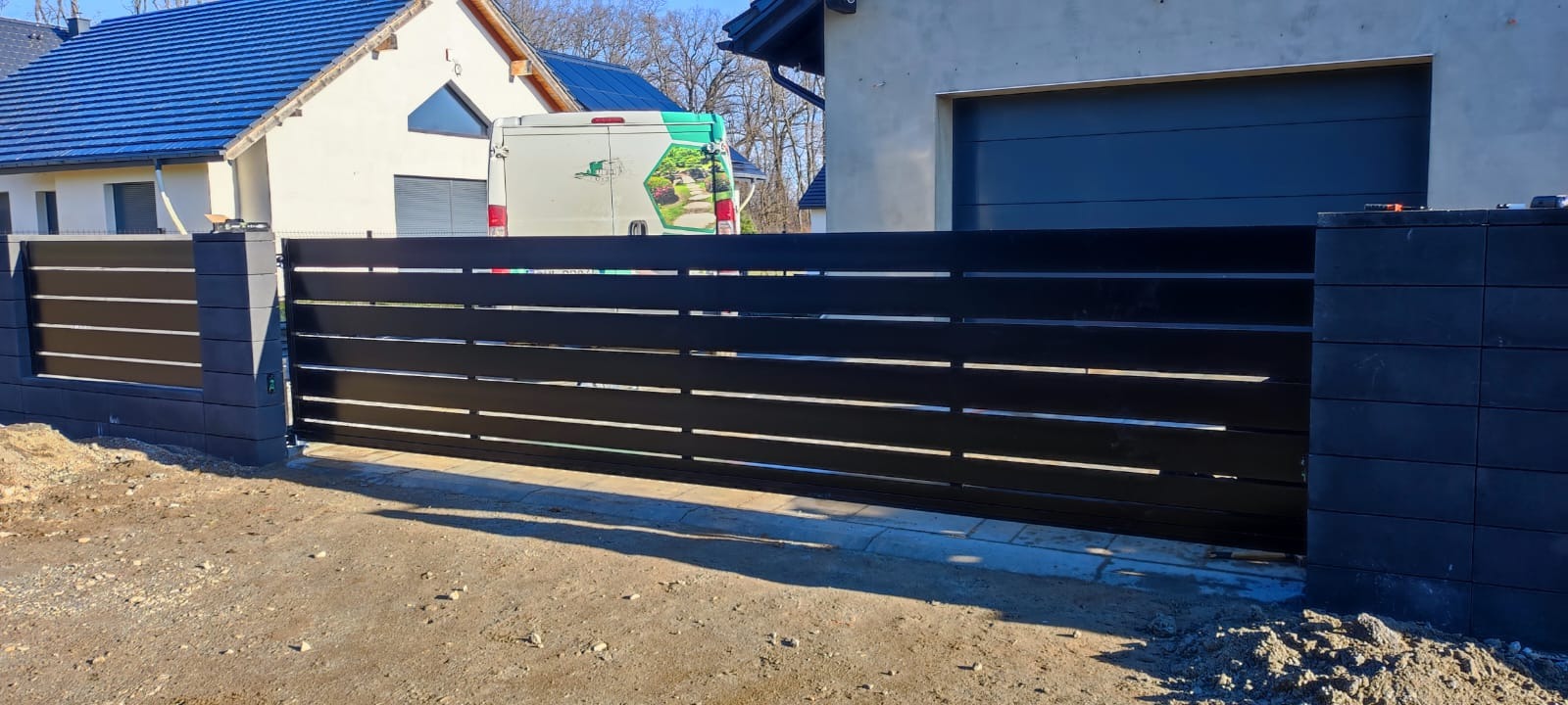 Nowoczesna brama wjazdowa przesuwna z poziomych profili metalowych w kolorze antracytowym, zamontowana na posesji z domem i garażem, widoczny samochód dostawczy z logotypem firmy w tle.