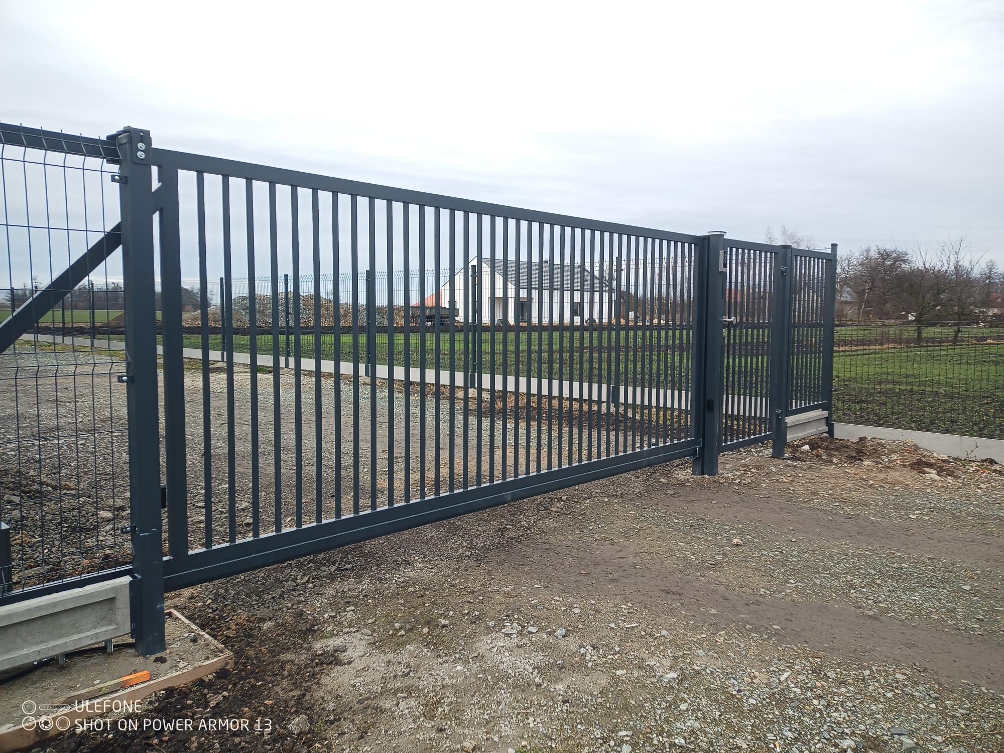 Szara, przesuwna brama wjazdowa z metalowymi prętami i panelem siatkowym, widoczny betonowy fundament i otaczające ogrodzenie panelowe, dom w tle.