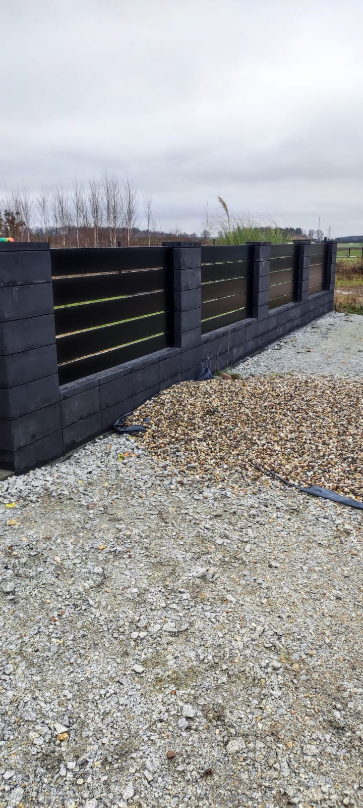 Nowoczesne ogrodzenie z czarnych paneli poziomych i szarych betonowych słupków, otoczone żwirem, na tle pochmurnego nieba i jesiennego krajobrazu.