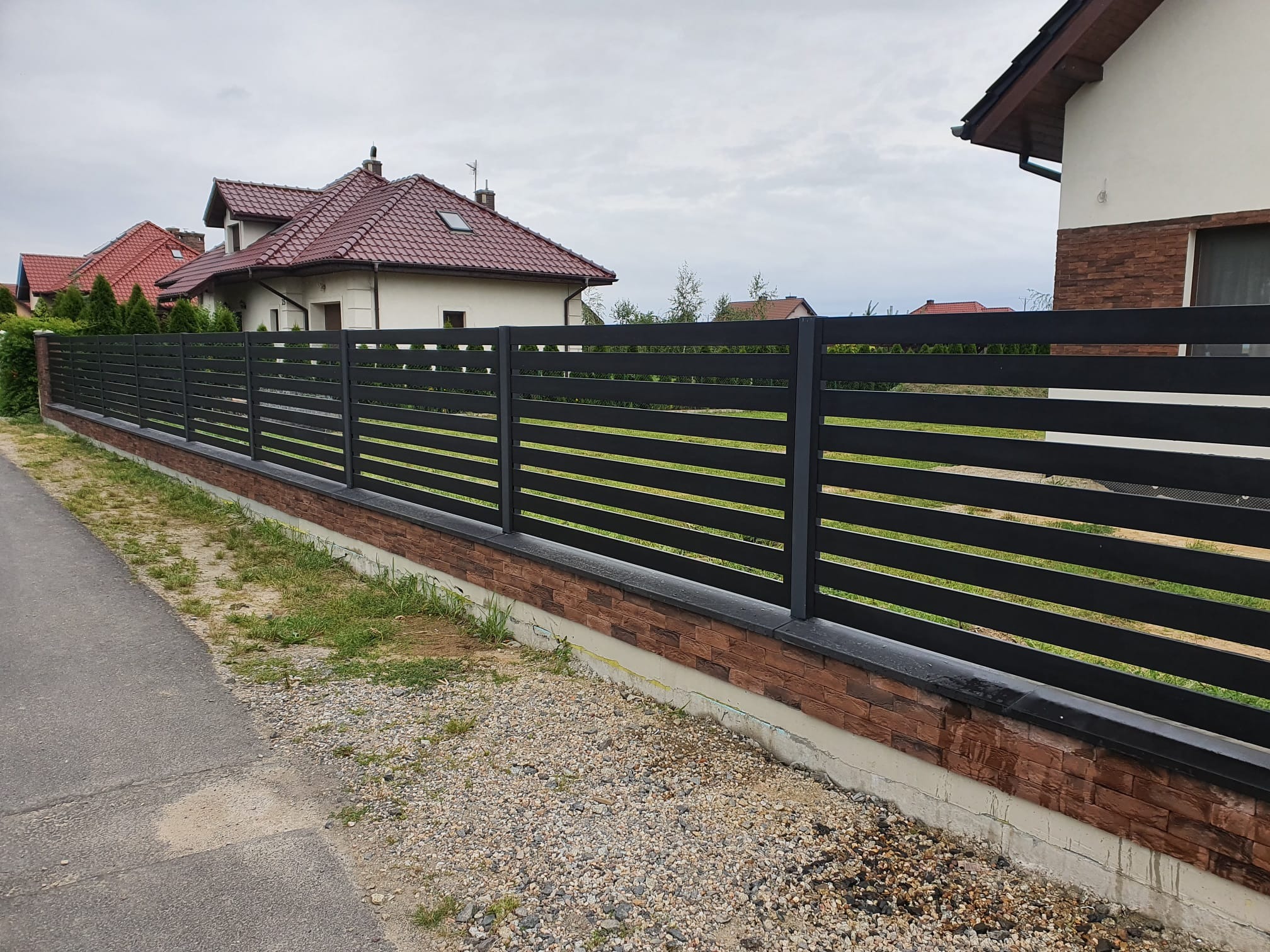 Nowoczesne, poziome ogrodzenie metalowe w kolorze grafitowym, osadzone na cokole z cegły klinkierowej. Widok z zewnątrz posesji, na tle domów jednorodzinnych.