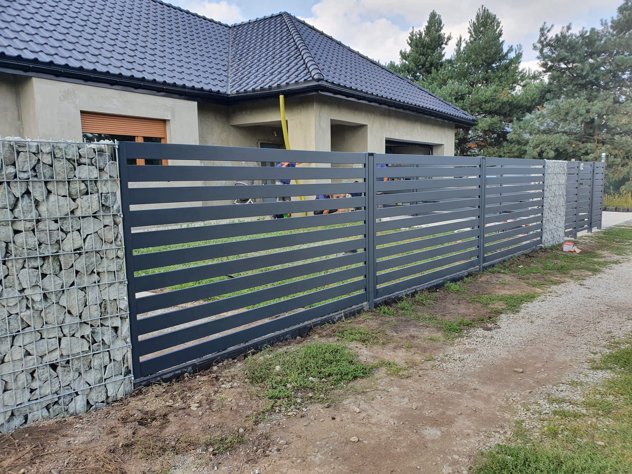 Nowoczesne ogrodzenie panelowe z elementami gabionowymi, szare, poziome panele na tle domu z ciemną dachówką i zieleni. Widoczne słupki i podmurówka.