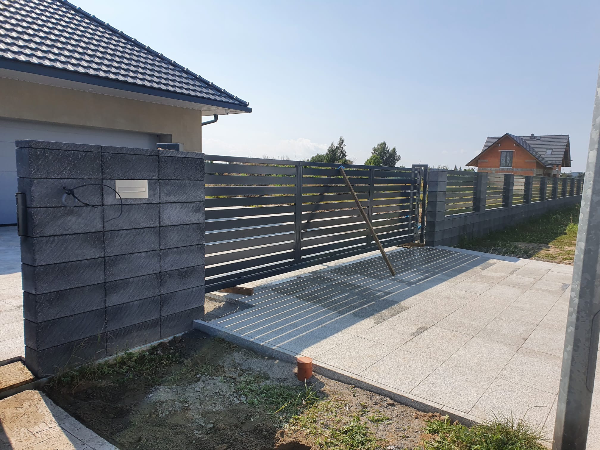 Nowoczesna brama przesuwna z poziomych profili, zamontowana na słupkach z szarej kostki łupanej. Widoczne płytki chodnikowe i fragment domu w tle.