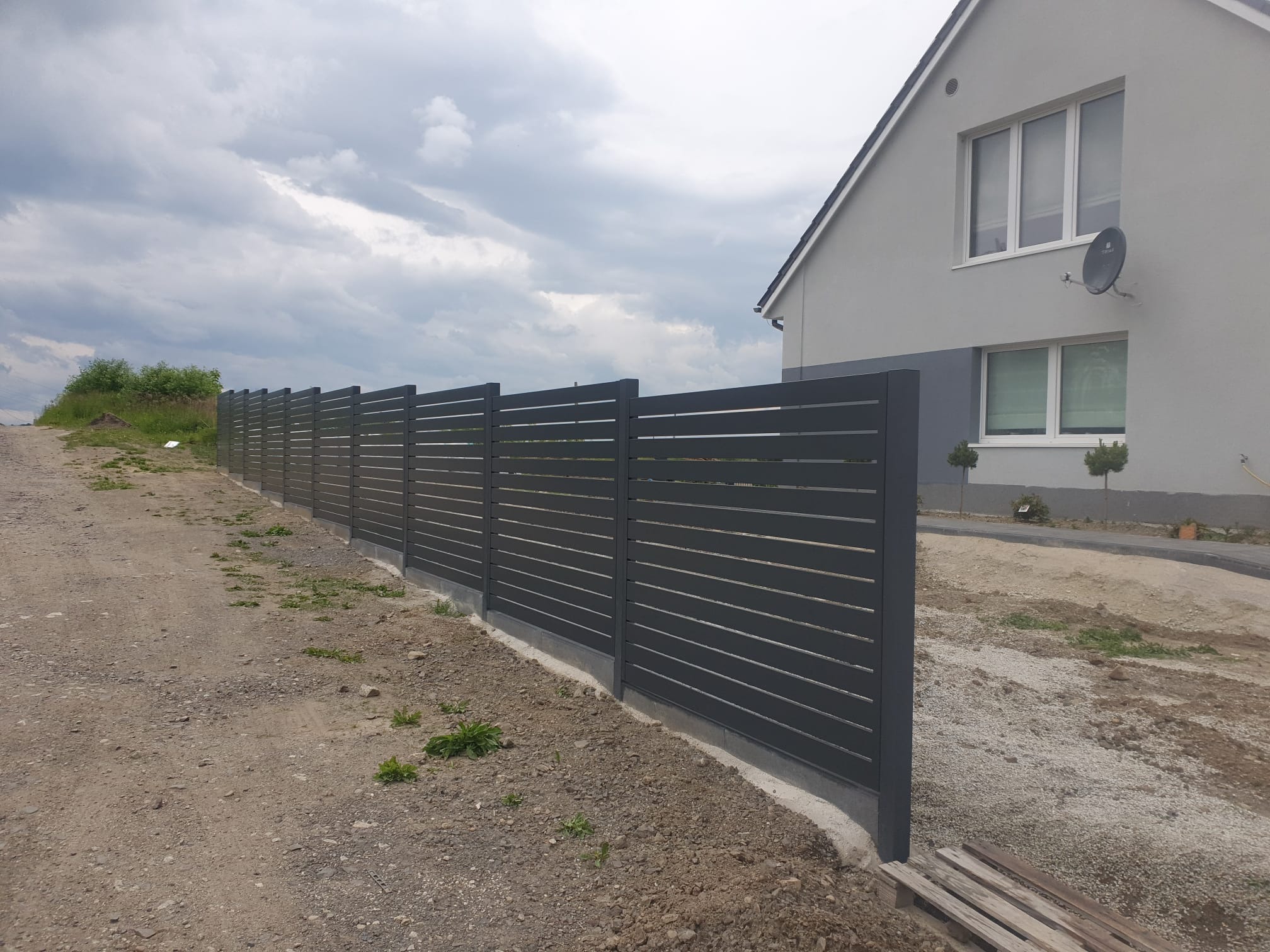 Nowoczesne, poziome, szare ogrodzenie panelowe z metalu, oddzielające posesję z domem od polnej drogi. Widok z boku, pochmurne niebo.