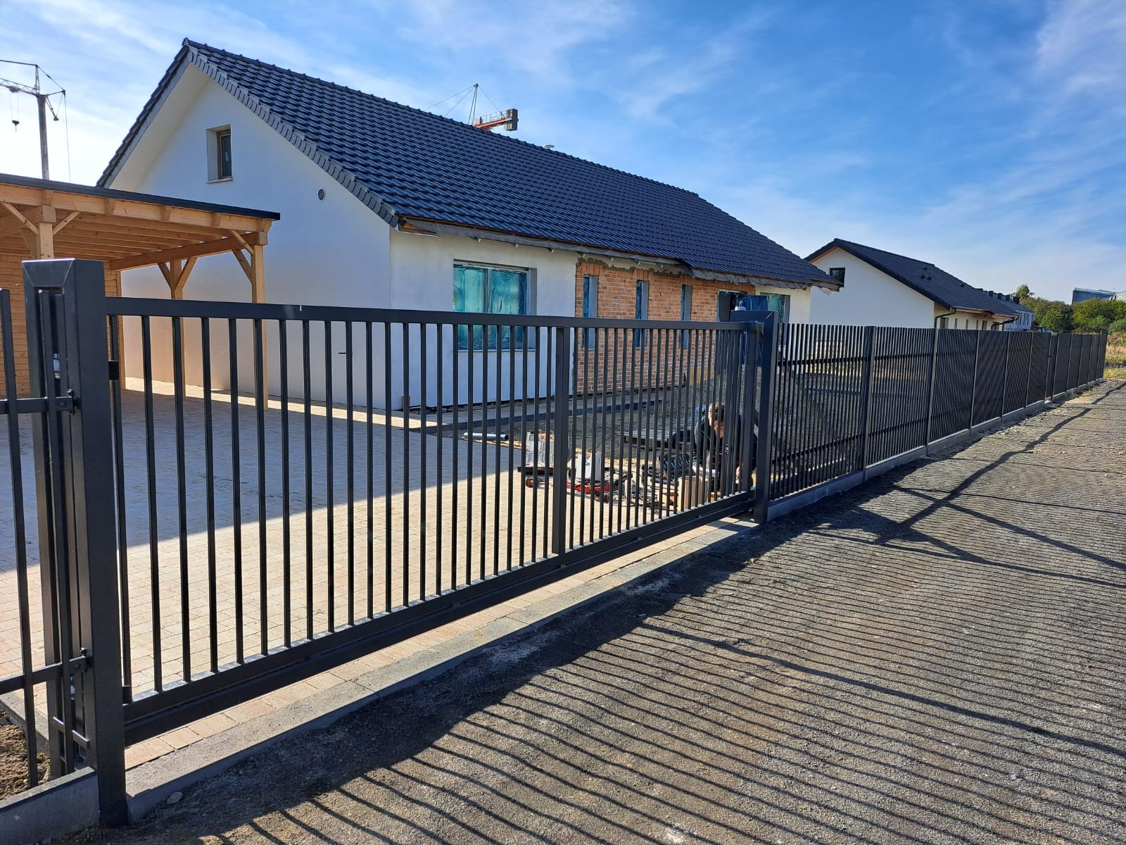 Nowoczesna, przesuwna brama wjazdowa z metalowymi sztachetami na tle domu jednorodzinnego w trakcie budowy. Widoczne narzędzia i osoba montująca.