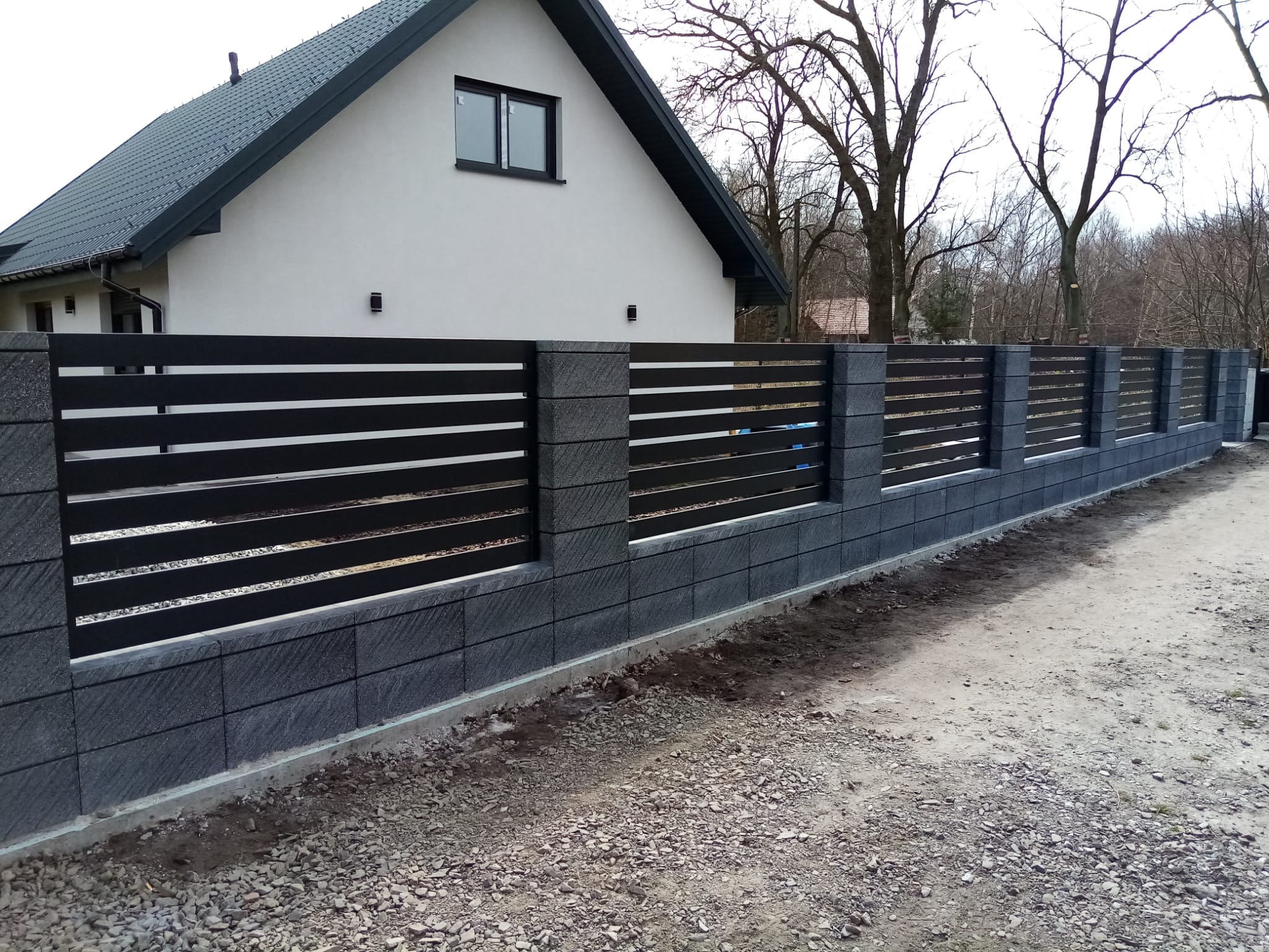 Nowoczesne ogrodzenie z szarej kostki łupanej i czarnych, poziomych paneli na tle domu z ciemnoszarą dachówką i drzew bez liści.