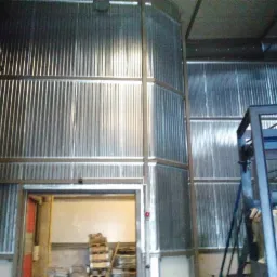 Wnętrze hali produkcyjnej z ścianami i sufitem wyłożonymi płytami warstwowymi, widoczne elementy konstrukcji stalowej oraz otwarte wrota magazynowe z paletami w środku.