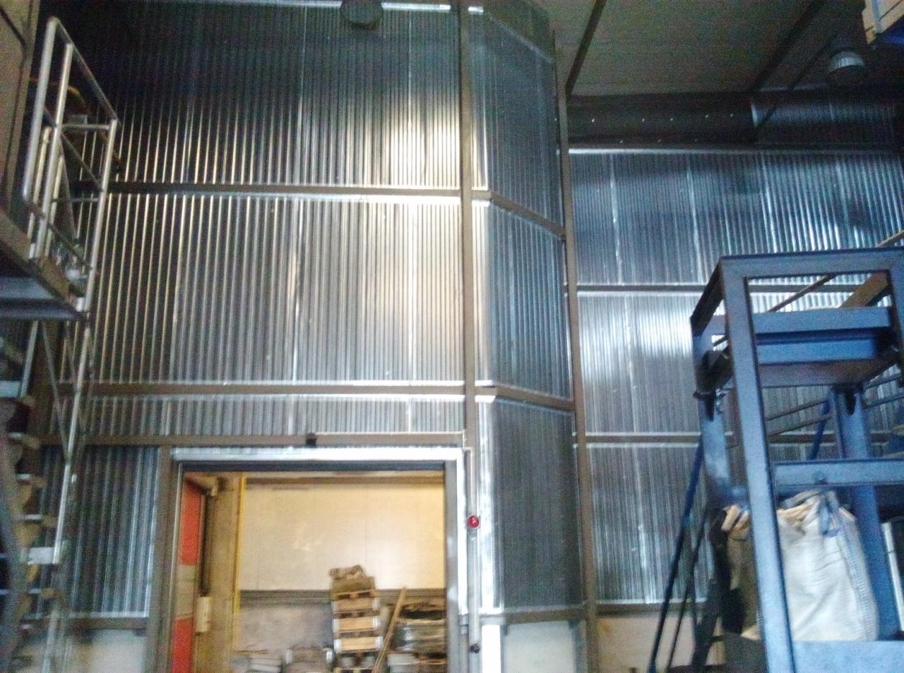 Wnętrze hali produkcyjnej z ścianami i sufitem wyłożonymi płytami warstwowymi, widoczne elementy konstrukcji stalowej oraz otwarte wrota magazynowe z paletami w środku.