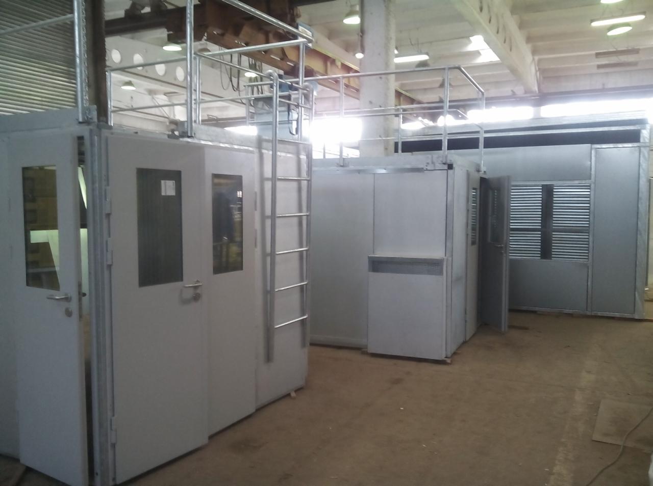 Wnętrze hali produkcyjnej z zamontowanymi kabinami z płyt warstwowych, wyposażonymi w drabinki i balustrady ochronne na górze, widoczne drzwi z oknami i kratki wentylacyjne.