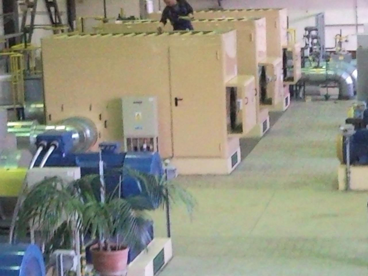 Rząd dużych, beżowych szaf przemysłowych w hali, widok z góry, z pracownikiem w kombinezonie na jednej z nich. Widoczne rury, kable i inne urządzenia. Na pierwszym planie roślina doniczkowa.