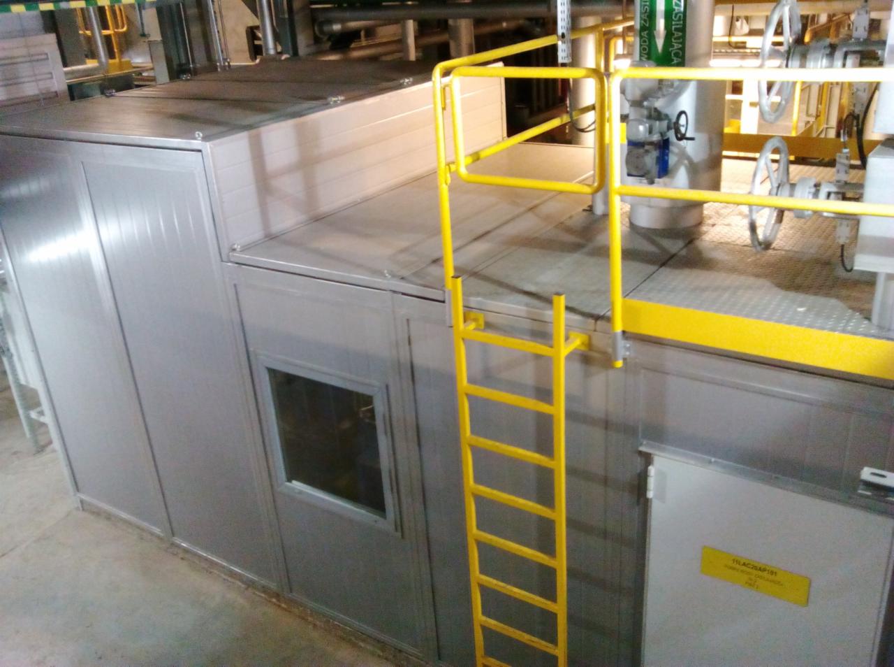 Szara konstrukcja z płyt warstwowych z żółtą drabinką, widoczne elementy instalacji przemysłowej w tle.