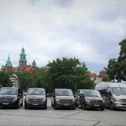 Flota ośmiu busów Mercedes-Benz w kolorach brązowym i srebrnym ustawionych rzędem na placu z widokiem na Wawel w Krakowie.
