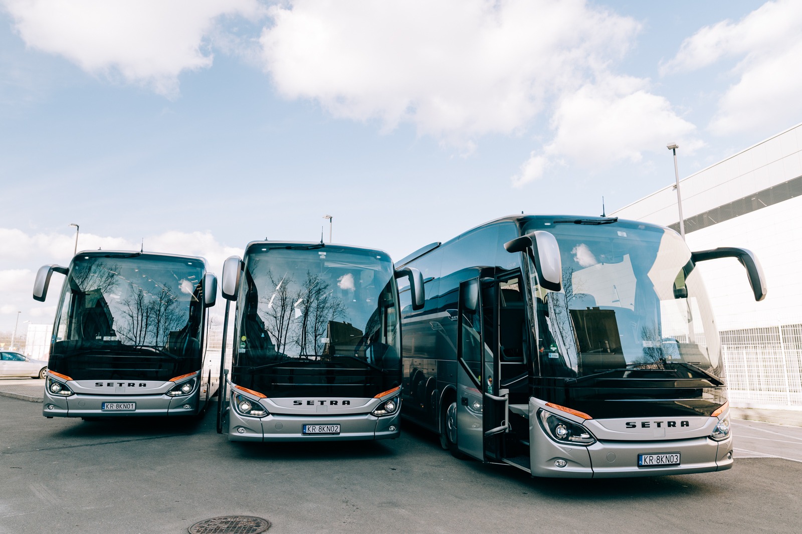 Trzy srebrne autobusy Setra zaparkowane w Krakowie z otwartymi drzwiami w jednym z nich, widoczne numery rejestracyjne KR 8KN01, KR 8KN02 i KR 8KN03, na tle błękitnego nieba z chmurami.