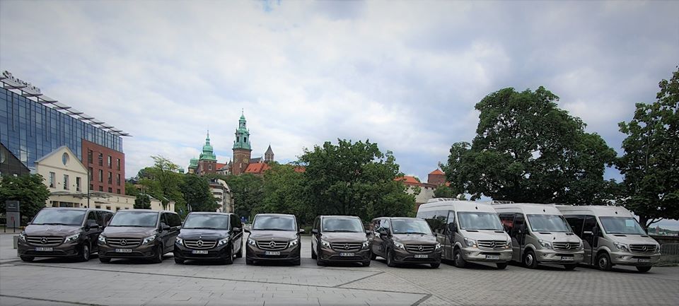 Flota ośmiu busów Mercedes-Benz w kolorach brązowym i srebrnym ustawionych rzędem na placu z widokiem na Wawel w Krakowie.