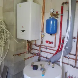 Biały piec grzewczy, niebieski zbiornik wyrównawczy i biały zasobnik ciepłej wody połączone miedzianą instalacją, widoczne również rury kanalizacyjne i rozdzielacz ogrzewania podłogowego.