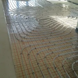 Białe rury ogrzewania podłogowego ułożone spiralnie na folii z czerwoną siatką, widoczne okna w tle i fragmenty ścian w surowym stanie.