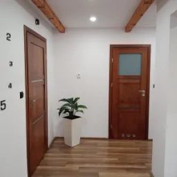 Wnętrze przedpokoju z drewnianymi drzwiami, belkami stropowymi, panelami podłogowymi imitującymi drewno, białymi ścianami z czarnymi cyframi 2-5, rośliną doniczkową w białej donicy i listwą...