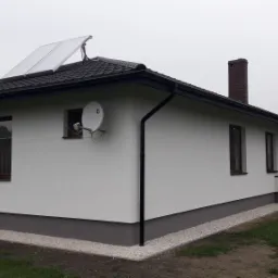 Biały, parterowy dom z ciemną dachówką i panelami słonecznymi, widoczna antena satelitarna na ścianie, szara podmurówka i białe kamyczki wzdłuż fundamentów.