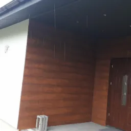 Elewacja budynku z drewnopodobną okładziną, brązowe drzwi wejściowe, biała ściana z oknem, dekoracyjna latarenka i donice z czerwonymi kwiatami przed wejściem.