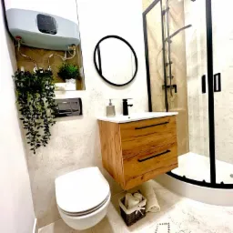 Nowoczesna łazienka z wiszącą szafką z drewnianymi frontami, okrągłym lustrem, kabiną prysznicową z czarnymi ramami i wiszącą toaletą. Nad toaletą zamontowany bojler i dekoracyjne rośliny.