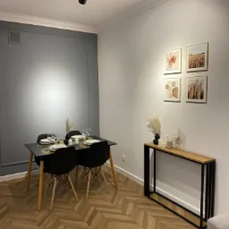 Nowoczesne wnętrze biurowe z drewnianą podłogą w jodełkę, czarnym stołem i krzesłami, szarą ścianą z dekoracyjną sztukaterią oraz minimalistyczną konsolą pod ścianą z galerią obrazków.